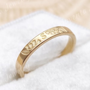 May include: A gold band ring featuring an engraved letter S and a delicate floral pattern of lily of the valley flowers. The ring has a polished finish and is positioned upright in a white velvet ring box with the text Juventa Jewelry visible.
