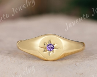 May include: A gold signet ring featuring a central starburst engraving with a small, round, purple gemstone set in the middle. The ring has a smooth, polished finish and a tapered band design, showcasing a clean and simple aesthetic.