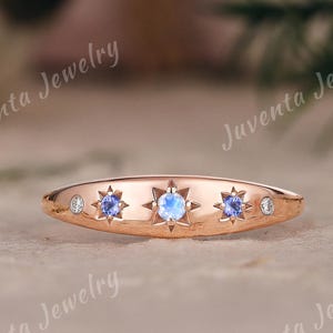 Op de afbeelding: Een roségouden ring met een centrale ronde maansteen, geflankeerd door twee kleine blauwe edelstenen en twee piepkleine heldere steentjes, allemaal gezet in stervormige gravures. De tekst Juventa Jewelry is zichtbaar op de achtergrond.