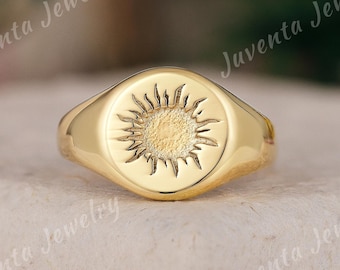 May include: A gold signet ring featuring a sunburst design. The ring has a polished finish and an oval face. The sunburst is etched into the center, with rays extending outwards. The ring is set against a neutral background.