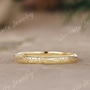 May include: A thin yellow gold band ring featuring an engraved floral pattern around the entire outer circumference. The surface of the metal is polished and reflective, showcasing intricate leaf and vine details etched into the gold surface.