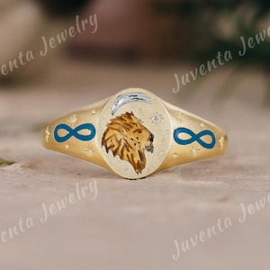 Puede incluir: Anillo dorado con un diseño de cabeza de león en el centro. El anillo presenta una luna creciente y estrellas, con símbolos de infinito azules a cada lado. El texto "Juventa Jewelry" es visible.