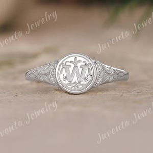 May include: A silver ring with a circular face featuring the letter "W" in a decorative font. The ring has a floral design on the band and small accent stones around the face. The ring is from Juventa Jewelry.