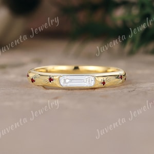 May include: A gold band ring with a rectangular, clear stone in the center. The ring is adorned with small red gemstones and star-like engravings. The ring is set on a neutral surface, with the words "Juventa Jewelry" in the background.