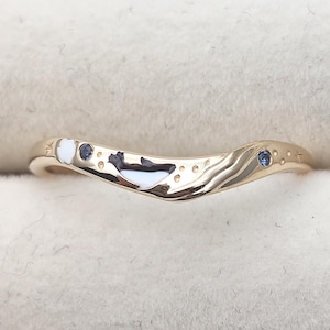 Könnte beinhalten: Ein goldfarbener, geschwungener Ring mit einer eingelegten schwarz-weißen Walsilhouette, einem kleinen weißen Akzent und zwei eingelassenen blauen Edelsteinen. Das Band ist mit eingravierten Stern- und Punktmustern auf der polierten Metalloberfläche verziert.