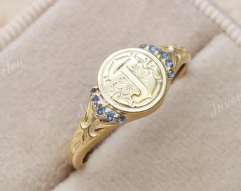 May include: A gold signet ring featuring a circular face engraved with the letter J and intricate floral patterns. The ring band is accented with small blue gemstones on both sides of the central setting, showcasing a polished metallic finish.