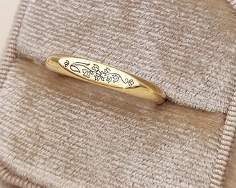 Könnte beinhalten: Ein polierter Goldring mit einem zarten, eingravierten Blumenmuster auf der Oberseite. Der Ring hat ein glattes, abgerundetes Profil und eine glänzende metallische Oberfläche, die feine Linien von kleinen Blumen und Blättern zeigt.
