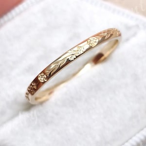 May include: A thin gold band ring featuring an engraved floral and leaf pattern along the outer surface. The ring is positioned within a white hexagonal velvet ring box. The text Juventa Jewelry is visible on the box surface.