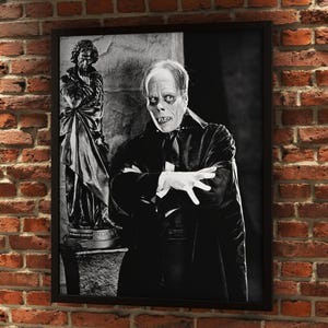 May include: Black and white framed print featuring a portrait of the Phantom of the Opera. The Phantom is in costume, with white makeup and a black cape. A statue is visible in the background. The print is displayed on a brick wall.