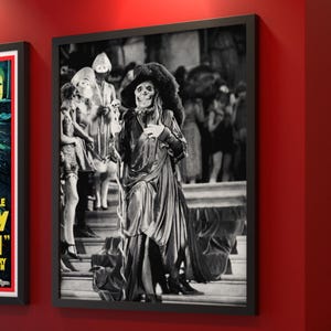 May include: Black and white framed print featuring a costumed figure with a skull face, holding a skull, and wearing a large hat. The figure is on a staircase with other figures in the background. The print is in a black frame.