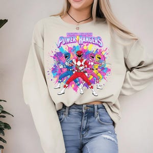 May include: Beige sweatshirt featuring a colorful graphic of the Mighty Morphin Power Rangers. The design includes the Power Rangers in their signature colors: red, blue, pink, and yellow, with the Power Rangers logo above them.