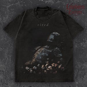 May include: Dark gray t-shirt featuring a distressed graphic of a knight in armor with the word "tired" above the image. The knight is surrounded by flowers. The shirt has short sleeves and a crew neck.