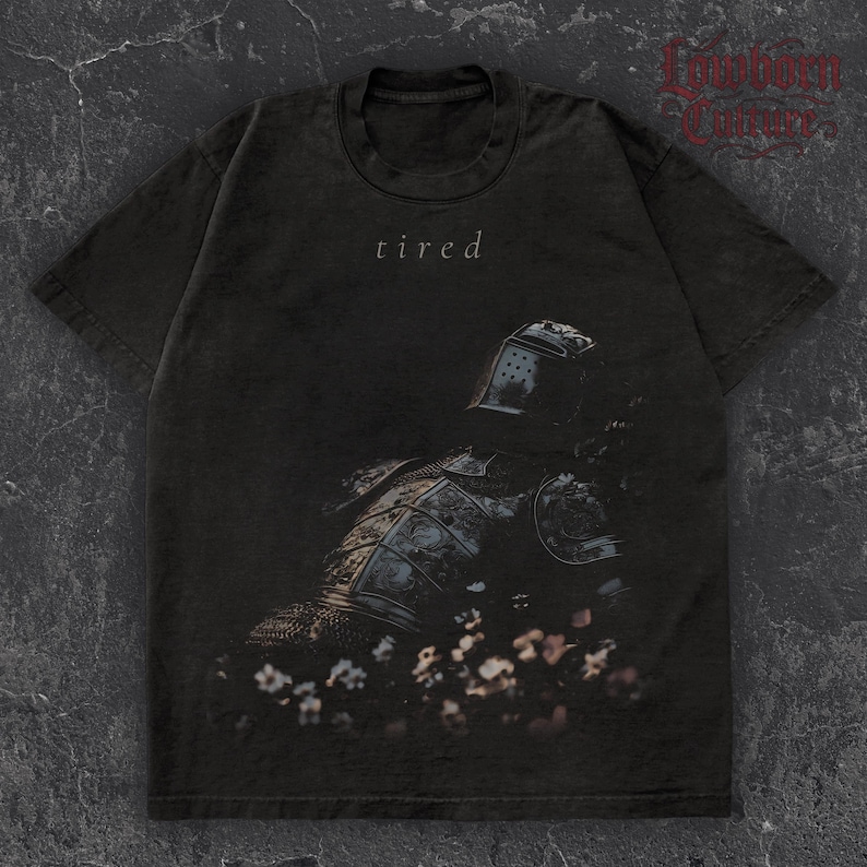 May include: Dark gray t-shirt featuring a distressed graphic of a knight in armor with the word "tired" above the image. The knight is surrounded by flowers. The shirt has short sleeves and a crew neck.