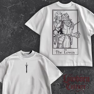 May include: White t-shirts with black graphic designs. One shirt features a sword, the other a tarot card illustration of two figures embracing, with the text "The Lovers". The shirts have a crew neck and short sleeves.
