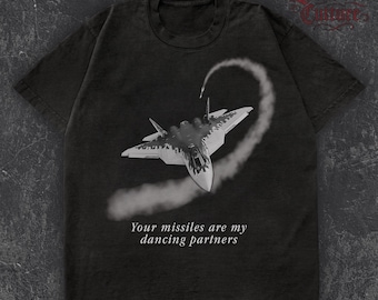 May include: Dark gray t-shirt featuring a fighter jet graphic with a vapor trail. The text "Your missiles are my dancing partners" is printed below the jet. The shirt has a crew neck and short sleeves.
