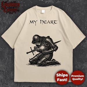 May include: Beige t-shirt featuring a black and white illustration of a kneeling knight with a sword, and the text "MY HEART". The shirt has a crew neck and short sleeves.