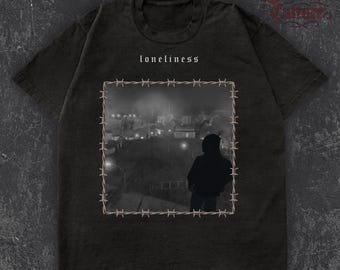 May include: Dark grey t-shirt with the word "loneliness" above a monochrome image of a person overlooking a town, framed by barbed wire. The shirt has a crew neck and short sleeves.