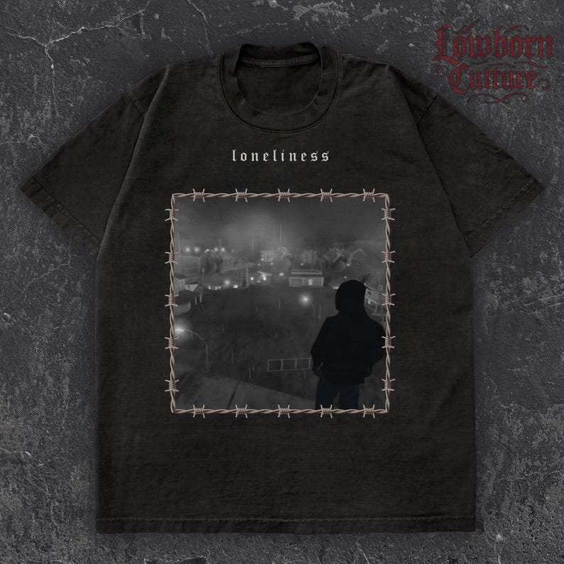 May include: Dark grey t-shirt with the word "loneliness" above a monochrome image of a person overlooking a town, framed by barbed wire. The shirt has a crew neck and short sleeves.