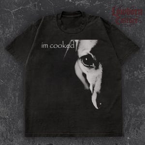 Puede incluir: Camiseta gris oscuro con la frase blanca "im cooked" y un gráfico en blanco y negro de un ojo. La camiseta tiene cuello redondo y mangas cortas. La tela parece suave y cómoda. Es una prenda informal.
