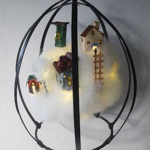 Op de afbeelding: Een decoratief hangornament met miniatuurhuisjes genesteld in een wolkenachtig materiaal, omsloten door een zwart metalen frame. De huisjes worden verlicht met warme lichten, waardoor een gezellige, grillige scène ontstaat. Een helder kristal hangt van bovenaf.