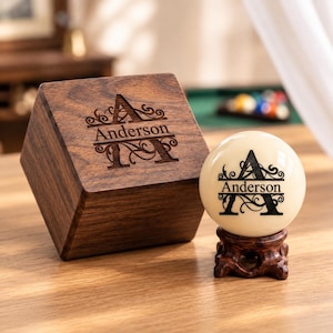 May include: A dark brown wooden box and a cream-coloured billiard ball resting on a small carved wooden stand. Both the box and the ball feature a laser-engraved design with a large letter A, decorative scrollwork, and the name Anderson.