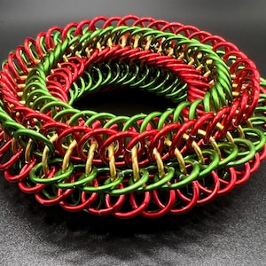 Puede incluir: Una pulsera circular de cota de malla construida con anillos metálicos entrelazados en colores rojo, verde y dorado. El diseño presenta un patrón tejido denso donde cada anillo individual se conecta con varios otros para formar una banda gruesa.