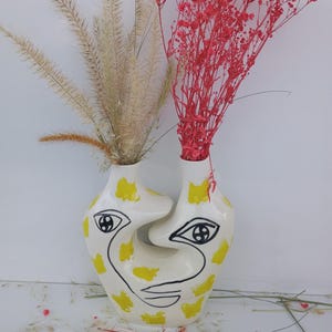 Peut inclure: Un vase en céramique blanc avec un design de visage, avec des contours noirs et des accents jaunes. Le vase contient des fleurs séchées, dont de l'herbe beige et du gypsophile rouge vif, créant un contraste saisissant de couleurs et de textures. Le vase a un design artistique unique.