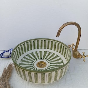 May include: A round ceramic sink with a green and white striped pattern. The sink has a brass drain and is paired with a brass tap. A small decorative dish and dried plant are also visible.