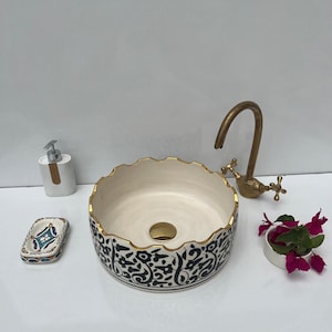 Peut inclure: Un lavabo en céramique avec un intérieur crème et un extérieur à motifs noirs et blancs. Le lavabo a un bord doré et est accompagné d'un robinet doré, d'un distributeur de savon, d'un porte-savon et d'un petit bol avec des fleurs.