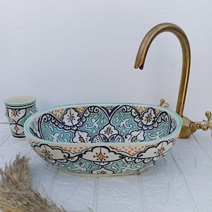 Puede incluir: Un lavabo ovalado de cerámica con un estampado floral turquesa y blanco. El lavabo está junto a una taza a juego y un grifo de latón. Las dimensiones del lavabo son de aproximadamente 46 cm de largo y 30 cm de ancho.