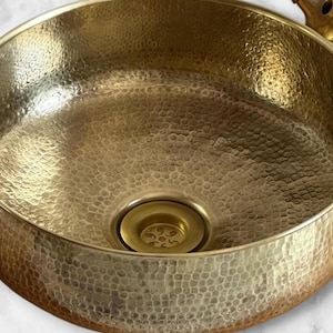 Puede incluir: Un lavabo redondo de latón martillado con una superficie texturizada. El lavabo tiene un desagüe central y un grifo de latón a juego. El diseño es vintage y decorativo, adecuado para un baño o cocina. El diámetro es de unos 30 cm.