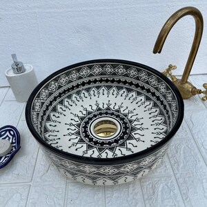 Peut inclure: Un lavabo rond en céramique décorative avec un motif géométrique noir et blanc. Le lavabo a un bord noir et un drain doré. Un robinet doré est visible en arrière-plan, ainsi qu'un distributeur de savon blanc.