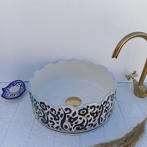 May include: A white ceramic vessel sink with a scalloped edge and a black floral pattern around the exterior. The sink has a gold-colored drain and is next to a brass faucet.