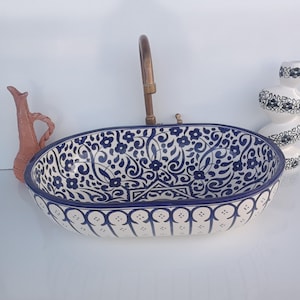 Peut inclure: Un évier ovale en céramique avec un motif floral bleu et blanc. Un robinet en bronze est monté au centre. Un petit vase en terre cuite et un vase décoratif blanc et noir sont de chaque côté de l'évier.