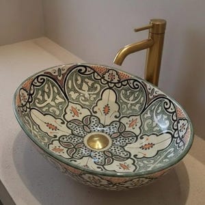 Peut inclure: Lavabo ovale en céramique avec un motif floral décoratif crème, vert, noir et orange. Le lavabo est associé à un robinet doré. Le lavabo est posé sur un comptoir de couleur claire.