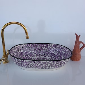 Peut inclure: Lavabo ovale en céramique avec un motif floral violet et blanc, souligné de noir. Un robinet doré et une petite cruche en terre cuite sont sur le côté. Un élément décoratif pour la salle de bain.