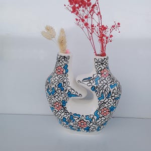 Peut inclure: Un vase en céramique blanc avec un design ouvert unique. Le vase est orné d'un motif floral bleu, rose et noir. Il contient des fleurs rouges et beiges séchées, créant un affichage décoratif.