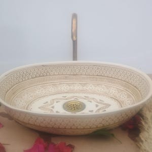Peut inclure: Évier ovale en céramique avec des motifs beiges et bruns complexes. L'évier présente un motif décoratif sur le bord et au centre, avec un drain et un robinet dorés. C'est un élément décoratif de salle de bain.