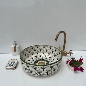 Peut inclure: Un lavabo rond en céramique avec un motif floral noir et blanc et un bord vert. Un robinet en laiton, un porte-savon et un distributeur sont présents, ainsi qu'une petite plante.