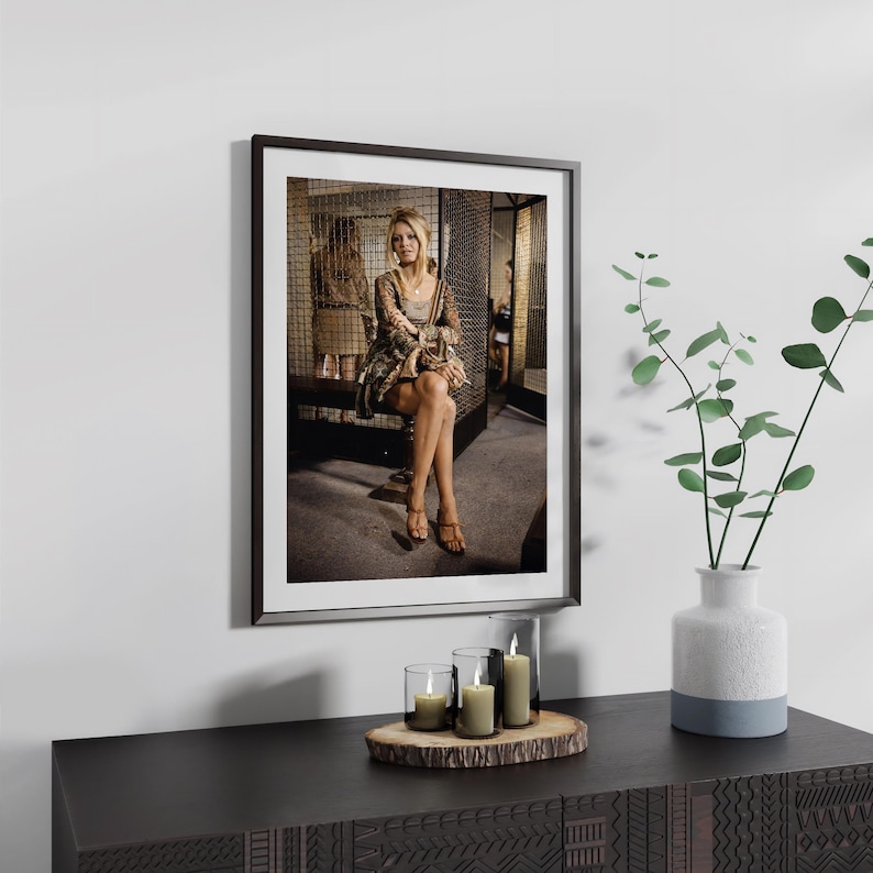 a framed photograph of a woman sitting on a bench, with a potted plant and candles on a wooden surface below the frame.
