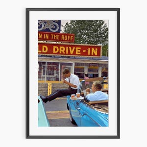 Puede incluir: Una impresión enmarcada de una escena vintage en un restaurante drive-in. Un hombre con camisa blanca y pantalón negro está sentado en el borde de un descapotable azul, comiendo. El letrero del drive-in dice "OLD DRIVE-IN" en rojo y amarillo.