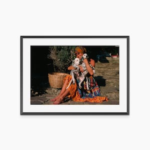 A woman in a colorful dress sits on a stone path, holding a small dog in her arms.