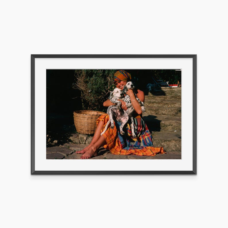 A woman in a colorful dress sits on a stone path, holding a small dog in her arms.