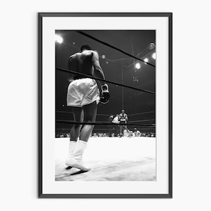 May include: Black and white framed photograph of a boxer in a boxing ring. The boxer is wearing white shorts, boots, and gloves, standing in the center of the ring. The image captures a moment of anticipation and focus, with the boxer's back to the viewer.
