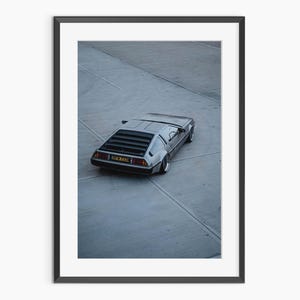 May include: Framed print featuring a silver DeLorean car, viewed from above, parked on a gray concrete surface. The car has distinctive gull-wing doors and a rear louvered window. The image is set in a black frame.