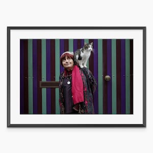 May include: Framed photograph featuring a woman with a cat perched on her shoulder. The woman is wearing a red scarf and a patterned coat. The background is a door with vertical stripes in purple, green, and blue.