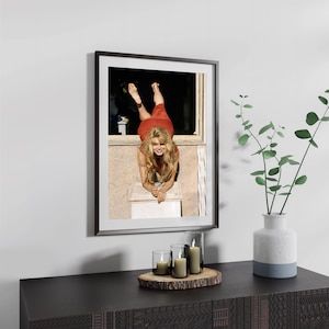 a framed photograph of a woman in a red dress, hanging on a wall above a wooden dresser with a vase of greenery and candles on it.