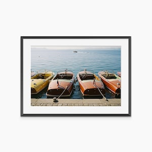 May include: A framed photograph featuring four wooden motorboats docked at a pier. The boats are various shades of brown and yellow, with red and white flags. The water is blue, and the sky is clear.