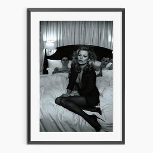 Può includere: Fotografia in bianco e nero incorniciata con una donna in blazer nero e tacchi seduta su un letto. Altre due persone sono parzialmente visibili sullo sfondo. La fotografia è in una cornice nera.