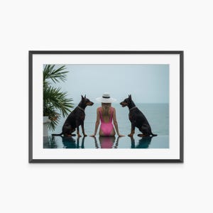 May include: A framed print featuring a woman in a pink swimsuit and a white hat, seated between two black Doberman Pinschers. The scene is set by a pool overlooking the ocean, with a palm tree visible on the left.